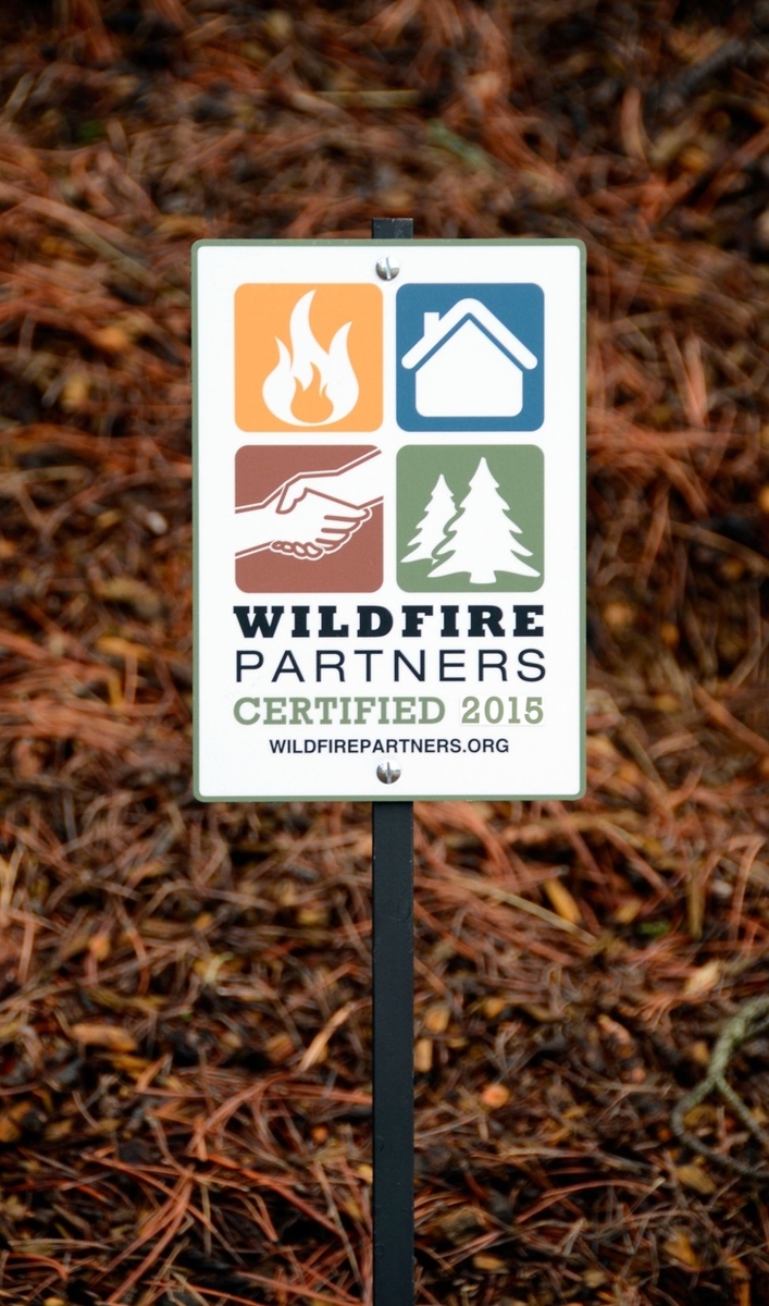 Sign reading “Wildfire Partners Certified 2015” posted in pine needles, with icons representing wildfire safety, home protection, neighborhood cooperation, and forested surroundings.